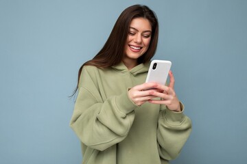 Smiling Beautiful young brunette woman wearing stylish green hoodie using mobile phone writing sms...