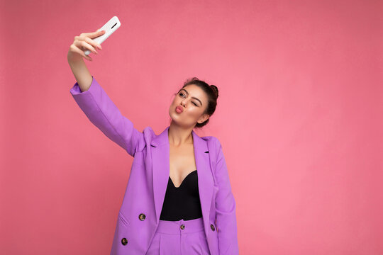 Photo of pretty young woman wearing purple suit taking selfie photo on the mobile phone isolated over pink background looking at phone camera