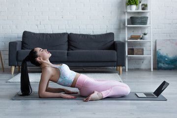 side view of woman practicing hero pose on yoga mat near laptop