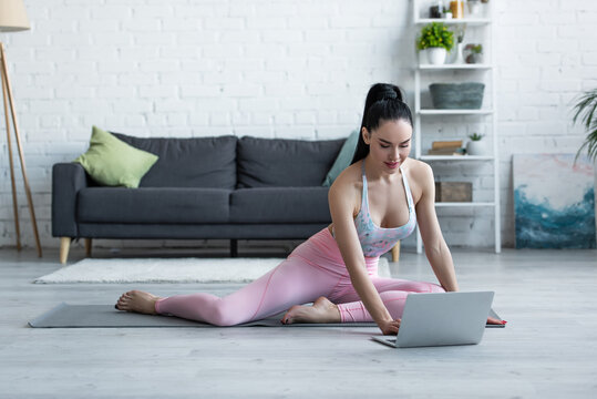 brunette woman in sportswear looking at laptop while sitting on yoga mat