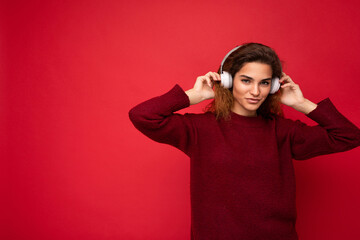 Beautiful happy smiling young brunette curly woman wearing dark red sweater isolated over red background wall wearing white bluetooth headphones listening to music and having fun looking at camera