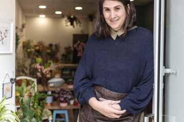 Portrait of a young cheerful woman working as florist standing at her own business shop entrance. Businesswoman concept. - obrazy, fototapety, plakaty