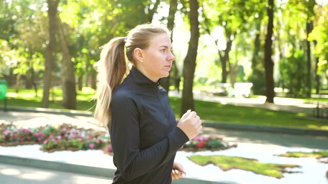Blonde woman with ponytail wearing black sportswear jogging in sunny park in slow motion, flower beds on background. Tracking shot athletic female training outside. Concept of healthy lifestyle