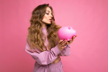 Portrait of doubtful thinking young beautiful attractive woman with blonde hair with sincere emotions wearing stylish pink hoodie isolated over pink background with copy space and holding pink piggy