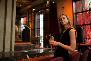 moody portraits of gorgeous blond girl with cocktail in fashion black outfit staying at the  bar in the club cafe or restaurant