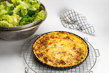 Freshly baked quiche  in black metal oven pie baking plate on a metal cooling rack with various vegetable and tablecloth on white background.