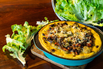 Freshly prepare homemade quiche in blue metal bowl on wooden board with green vegetables on wooden table.