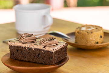 Mini chocolate cream sponge cake in wooden plate and piece of prune sponge cake in wooden dish with a cup of coffee and metal spoon on wooden table