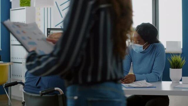 Invalid paralysed handicapped office manager explaining to black colleague financial startegy pointing graphs on clipboard wearing face masks against covid19 pandemic in new normal company