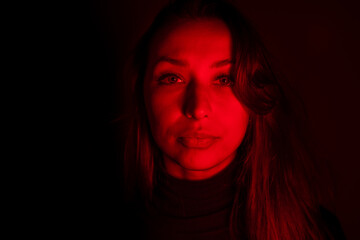 Young pretty woman looks at camera in contrast red light. Sensual female person posing in dark studio with red neon lightning. Portrait of calm woman, abuse addiction, depression, imprisonment concept