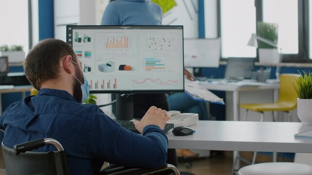 Diverse businesspeople using computer desktop, showing new startup idea, sitting at desk in new normal company office wearing protection mask, team leader with disabilities sitting in wheelchair