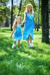 Mother and daughter are outdoors in a sunny park. Daughter leads mom by the hand.