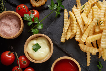 Homemade baked potato fries with mayonnaise, tomato sauce and rosemary on paper over black stone background. . tasty french fries, unhealthy food.