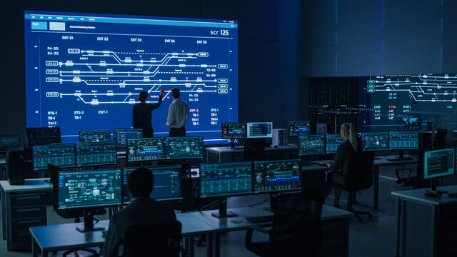 Project Manager and Computer Science Engineer Talking while Using Big Screen Display Showing Infrastructure Infographics and Data.Telecommunications Company System Control and Monitoring Room