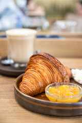 Ceramic plate with fresh crispy croissant, orange jam and butter with a glass cup of cappuccino on wooden table in cafe.