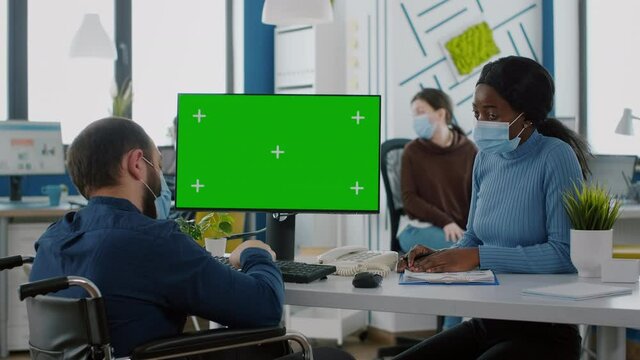 Invalid paralysed company manager with face mask typing on computer with green screen choma key mockup display discussing with african employee. New normal business office respecting social distance