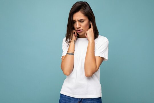 Portrait of sad upset young beautiful brunette woman with sincere emotions wearing casual white t-shirt for mockup isolated on blue background with empty space and covering ears with fingers