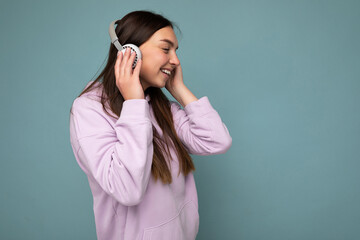 Beautiful happy smiling young brunette female person wearing light purple hoodie isolated over blue background wall wearing white bluetooth headsets listening to cool music and looking to the side