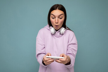 Beautiful surprised young brunette woman wearing purple hoodie isolated on blue background holding...