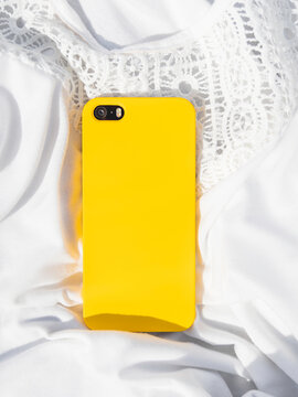 Smartphone in bright yellow case on white crumpled t-shirt with laces. Light textile background with vibrant cellular phone. Wireless technological device on summer outfit.