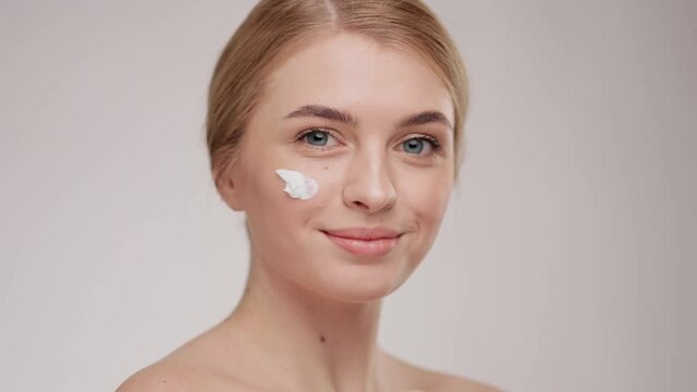 Young blonde hair woman takes care of her skin and smiling on camera. Closeup portrait young woman uses creams of her face. Fresh moisturized skin. Enjoy process smiles caring about perfect skin.