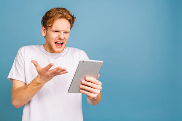 Happy shocked amazed young bearded red-haired man in casual using tablet and standing isolated over...