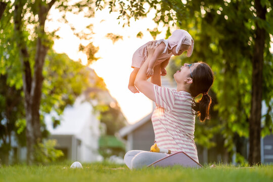 A mother was raising her little baby up in the air to play with her child on the green grass under the village tree. Backlit photos during the golden hour.
