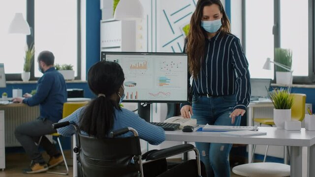 Company manager with medical mask against coronavirus explaining to african paralysed colleague project plan sitting at desk in new normal start up business office. Multiethnic business team working