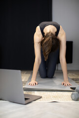 Fototapeta na wymiar Beautiful young woman dressed in black sportswear enjoying yoga indoors. Yogi girl working out on hands and knees in Table pose Listening to online mentor in laptop, youtube yoga courses for beginners