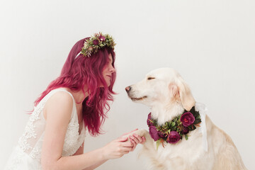 Young woman in a wedding dress with pink hair holds the paw of a golden retriever in a flower wreath on a white background
