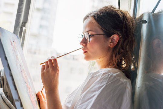 Side view of a pretty female artist painting on canvas in her art studio sitting next to the window. A woman painter with eyeglasses painting with oil searching for imagination in the workshop.