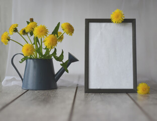 On a wooden table, a bouquet of yellow dandelions in a blue watering can and a photo frame. In the...
