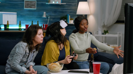 Black woman experiencing virtual reality headset winning video games using gaming joystick. Mixed race group of friends hanging out together drinking beer having fun late at night in living room