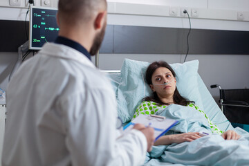 Specialist oncologist physician writing disease examination on clipboard monitoring sickness symptom working in hospital ward. Sick patient resting in bed discussing illness treatment