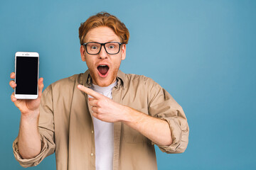 Happy winner! Young handsome shocked amazed man showing smartphone screen isolated over blue...