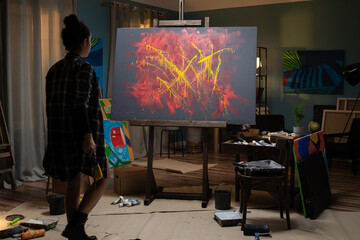 A young painter stands in her studio facing a painting and evaluates her project, a work that is an abstraction. The artist holds a paintbrush soiled with paint in her hand