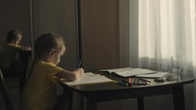 Side view of little preschool girl sitting at the table in her bedroom in front of window and does homework writing in practice book