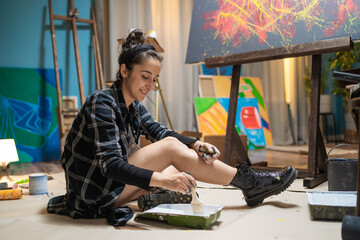 Artist in a checkered shirt and clogs is sitting on the floor secured with cardboard, among paintings, holding a brush in her hand. Her hands are dirty, but she is smiling and happy with her work