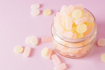 Chewing sugary citrus gummy candies in glass jar on pastel pink background. Top view, selective focus.