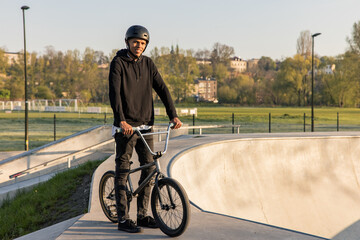 A young, smiling boy is wearing a helmet on his head and standing on a ramp with a bike to do tricks.