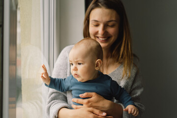 Mother playing with baby son at home near the window. Happy infant and mom. Isolation during quarantine on Mother's day