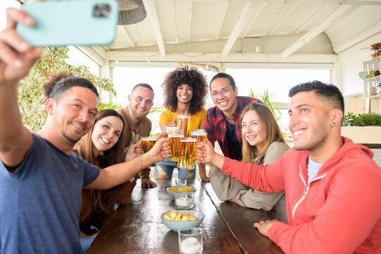 Multiracial friends taking selfie while toasting beer at restaurant - Young people having fun with drinks - Lifestyle concept about boys and girls make a party in happy hour at bar