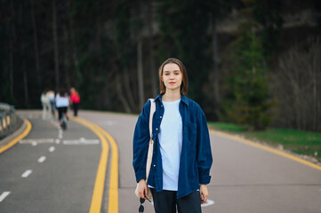 Attractive woman in a denim shirt stands on a mountain walking road on a background of coniferous trees and poses for the camera. Portrait of a tourist on a walk
