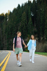 Obraz premium Couple of stylish tourists spend time together in the mountains, holding hands walking on the road in the woods and smiling.