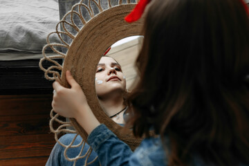 A girl with brown hair looks at her reflection in the mirror. Serious look. On the face are smears of paint and on the head is a red bandana tied with a bow. She is wearing a blue jumpsuit. Portrait