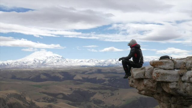 a male tourist traveler on a hill overlooking the mountain range of the Caucasus mountains Elbrus sits drinking tea from a thermos