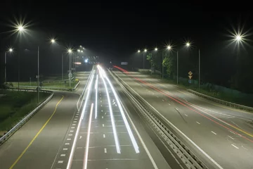Gordijnen A country highway at night with long streaks of light from the headlights of passing cars. There are streetlights at the edges of the road © VeNN