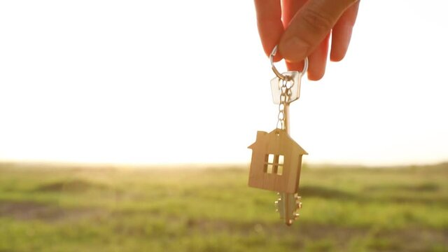 House key with keychain in hand. Background of the sky, sunlight and field. Dream of home, building a cottage in the countryside, plan and project design, farm, moving to a new home. Copy space