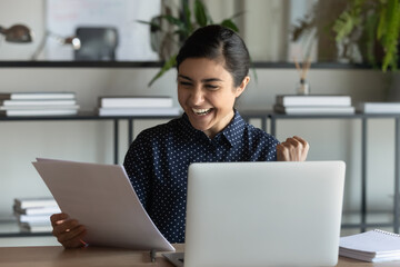 Overjoyed millennial Indian female employee triumph read good news work online on laptop in office....