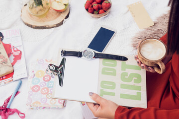 Female's hand is holding a clipboard with lettering 'to do list" and a black watch on it with blurred strawberries, apple, magazine in the background.